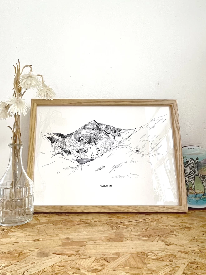 A print of a line drawing of Snowdon Mountain in Snowdonia National Park, in a frame next to some flowers, a rock and a candle