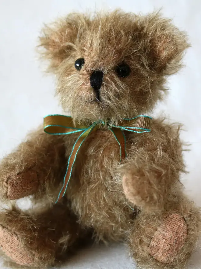 Small handmade brown teddy bear with a ribbon bow sitting against a plain background.