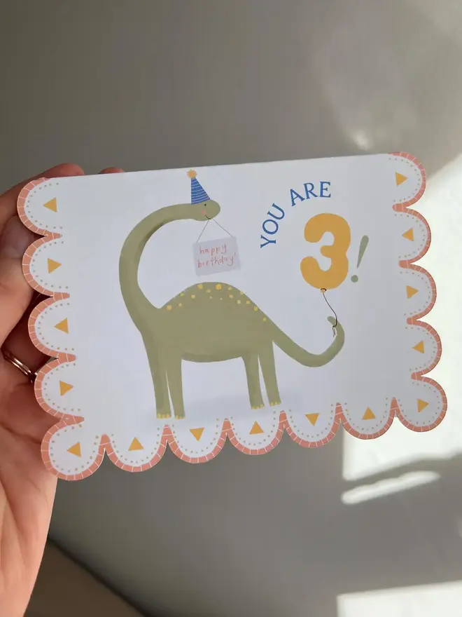 illustrated dinosaur 3rd birthday card. scallop edged birthday card illustrated with green stegosaurus.