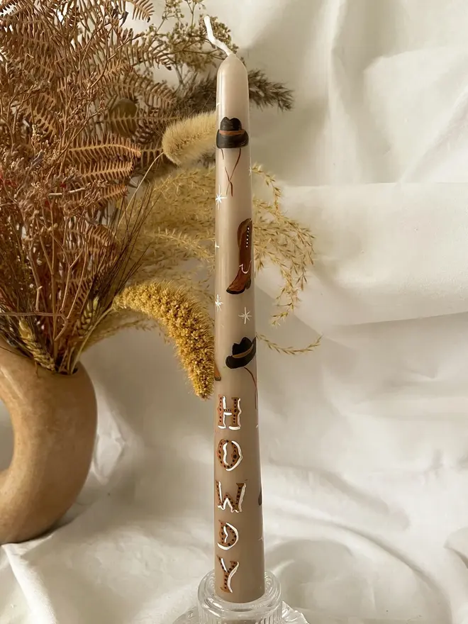'Howdy' Personalised Hand Painted Candle, a candle in a glass holder on a white surface accompanied by a plant. It's design can be seen wrapping around the stick. 