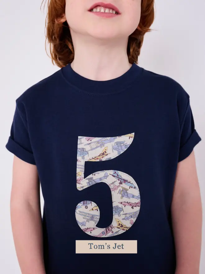 a 5 year old boy wearing a navy birthday t-shirt with a number 5 cut from liberty vintage plane print on the front