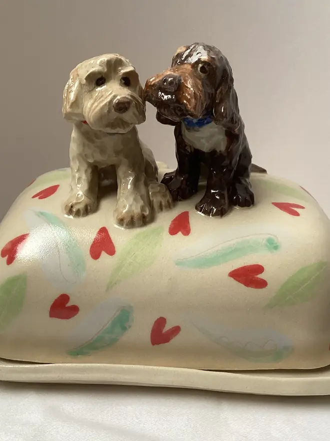 Two dogs looking at each other on a butter dish. Base is cream with red hearts and green and blue paint flicks.