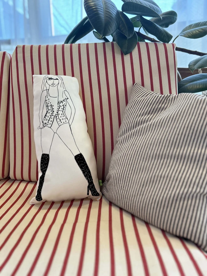 Illustrated black and white screen printed cushion of singer Taylor Swift on a striped sofa beside some plants.