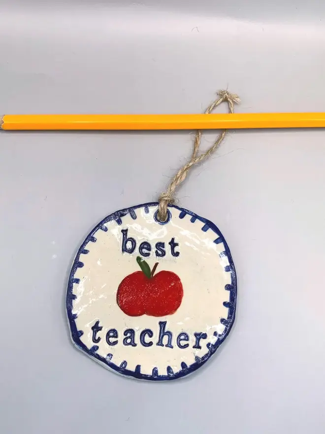 'Best Teacher' Handcrafted Ceramic Ornament, a ceramic apple ornament sitting on a plain white surface hanging from a pencil. 