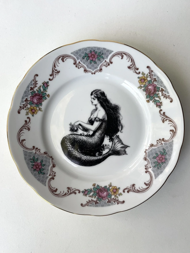 a vintage plate with an ornate border has a black and white vintage illustration of a mermaid in the centre