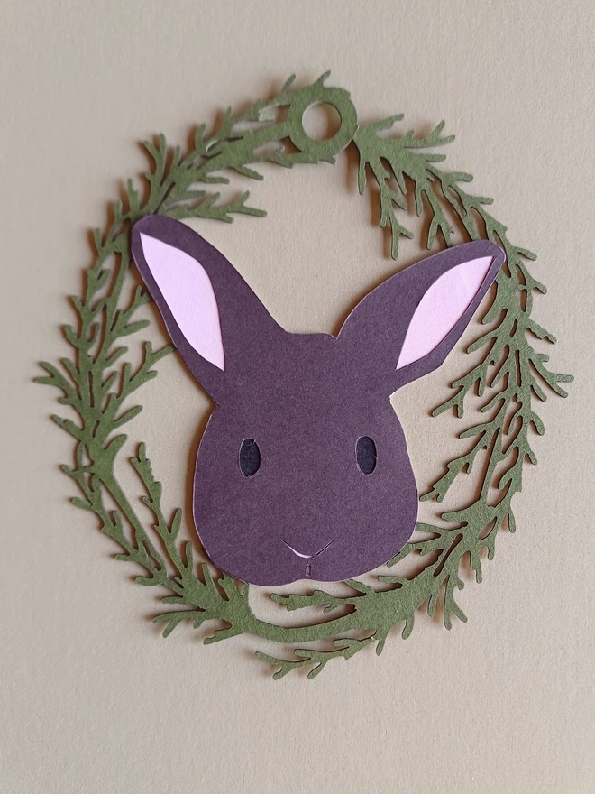 Black Bunny Easter Tree Decoration. Dark brown bunny face with black eyes and pale pink ears inside olive green foliage wreath oval