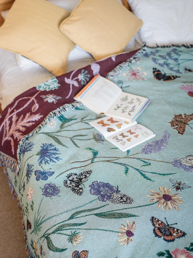 'Pollination Meadow' Recycled Cotton Blanket seen on a bed with yellow pillows.