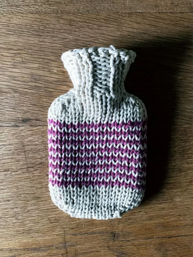 Mini Hot Water Bottle in hand knit string cover with purple stripe on an oak table