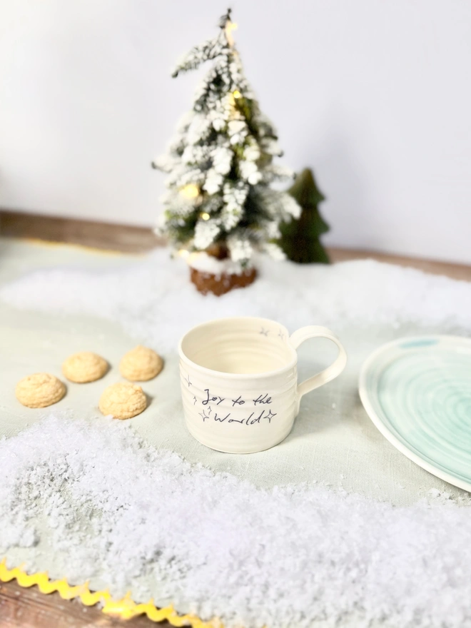 cream hand thrown porcelain mug with the words 'Joy to the World' printed onto the side with pretty line drawn stars. Sitting on a tabletop with snow, a mini lit Christmas tree and some biscuits