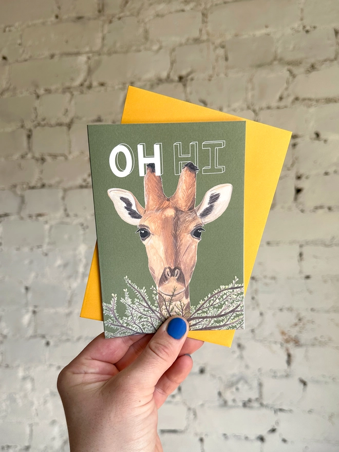  a green greetings card featuring an illustration of a giraffe peering over a bush with a deep yellow envelope