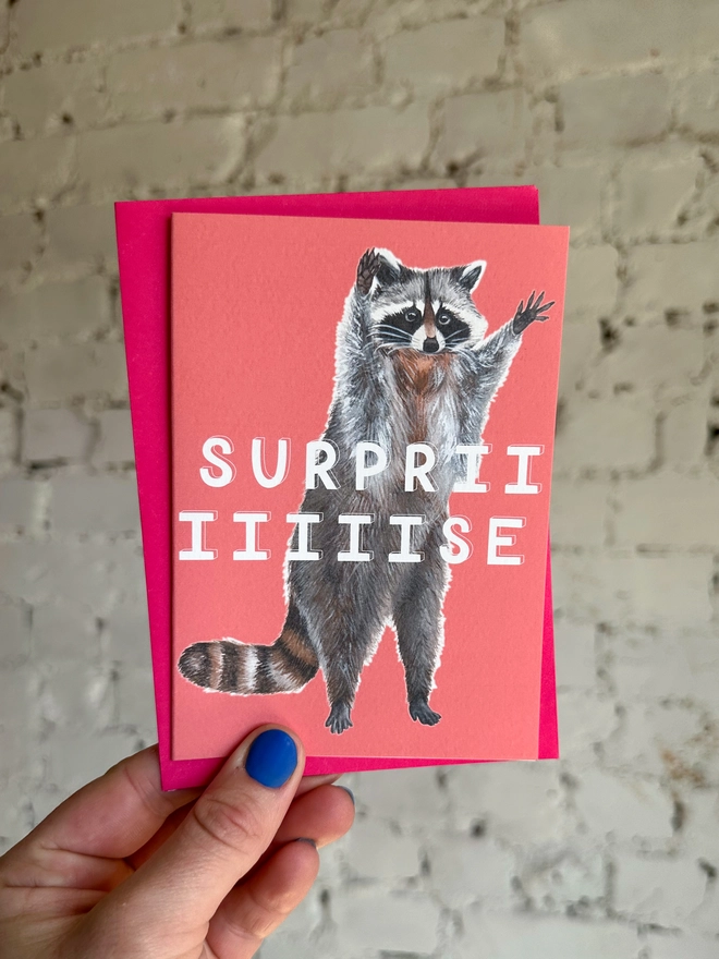 a bright coral pink greetings card featuring an illustration of a raccoon with its hands in the air with the text “surpriiiiise” with a neon pink envelope