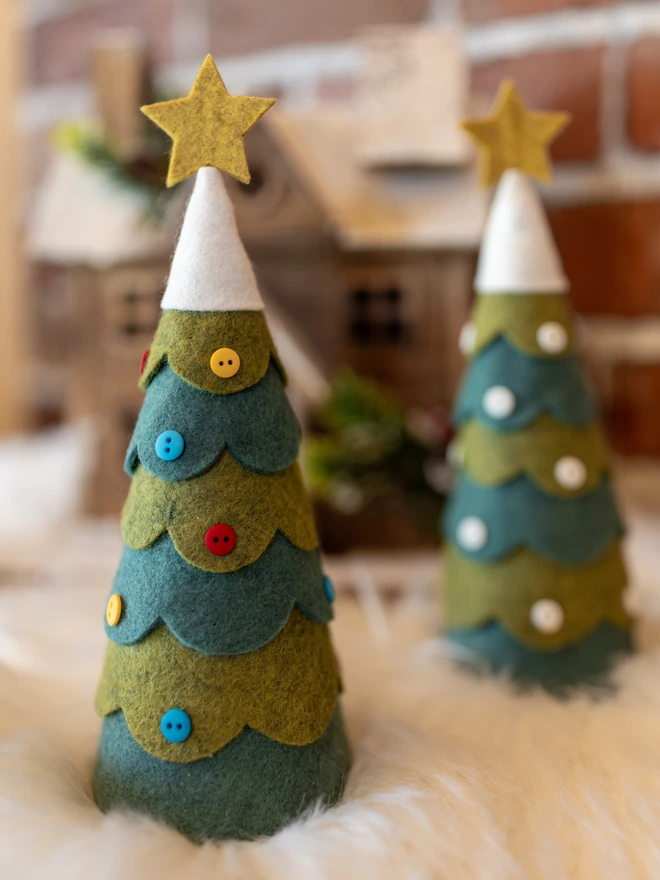 A pair of felt Christmas tree decorations. Each is decorated with buttons, one with all white, the other with red, yellow and blue. The tree has a snowy felt top adorned with a star.