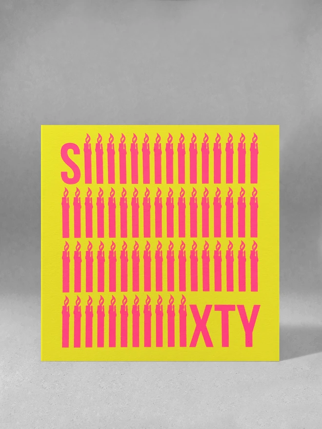 The word Sixty screen printed in neon pink ink across a bright yellow square card. To emphasise the sixty, the letter i is shaped like a simple lit candle and is repeated sixty times in four rows. The card is stood square on in a light grey studio set
