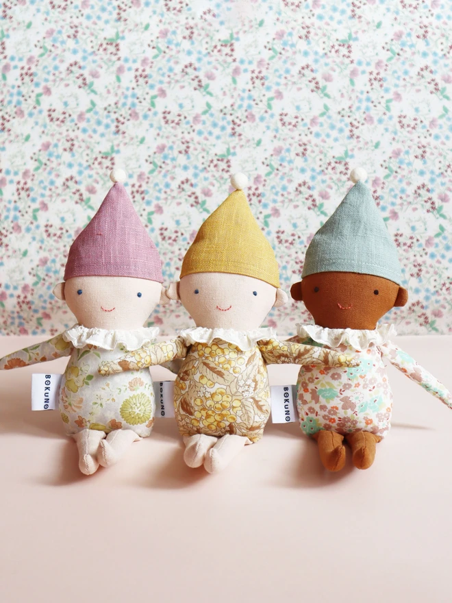 Three handmade Spring Elf Dolls are sitting side by side against a soft pastel background. Each doll has a floral print body, pastel-coloured hat with a pompom, and a ruffled white collar. They feature different skin tones and gentle smiles, radiating charm and warmth.