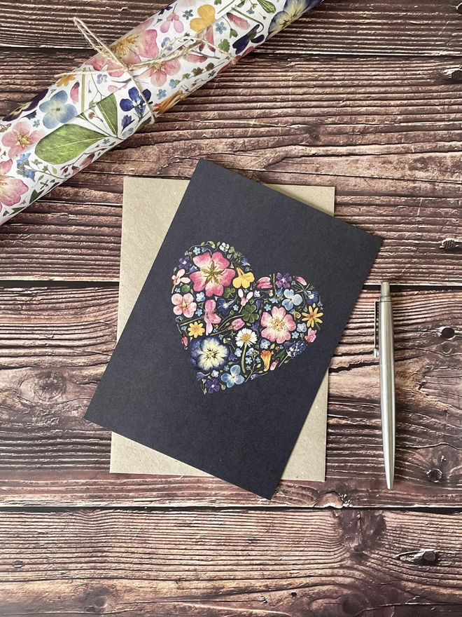 Floral Greetings Card with Pressed Flower Heart Design and Brown Kraft Envelope - Dark Brown Wooden Background - Silver Parker Pen - Rolled Pressed Flower Wrapping Paper