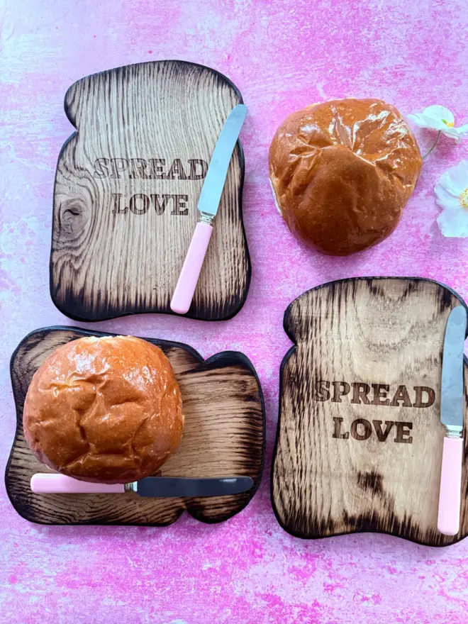 Three toast shaped serving boards with the message spread love engraved with pink vintage butter knives on pink background