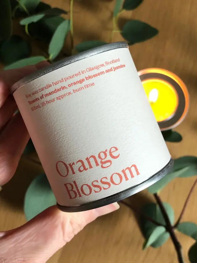 Orange Blossom Candle, held in a hand against a wooden and plant background. 