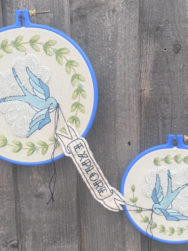 Two embroidery hoops joined by a banner with the word explore stitched onto it two bluebirds swallows birds in front of white beaded clouds and wreaths of green leaves 