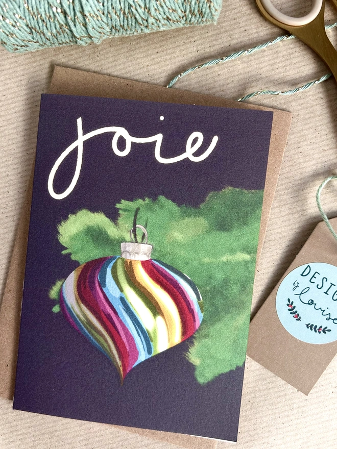 An illustration of a striped rainbow coloured Christmas bauble hanging from the branch of a Christmas tree on a deep purple background. The French word 'joie' is written above in white cursive handwriting.