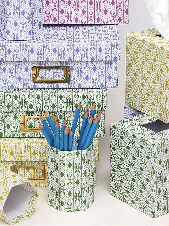 Tijan Hexagonal Brush Pot, a colourful patterned pencil pot sitting on a white surface accompanied by some folders. It is filled by some pencils. 