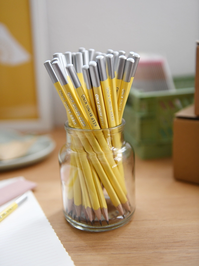 A jar full of yellow pencils each with the words "Keep Being Kind" along the side stands on a wooden desk with various stationery items around it.