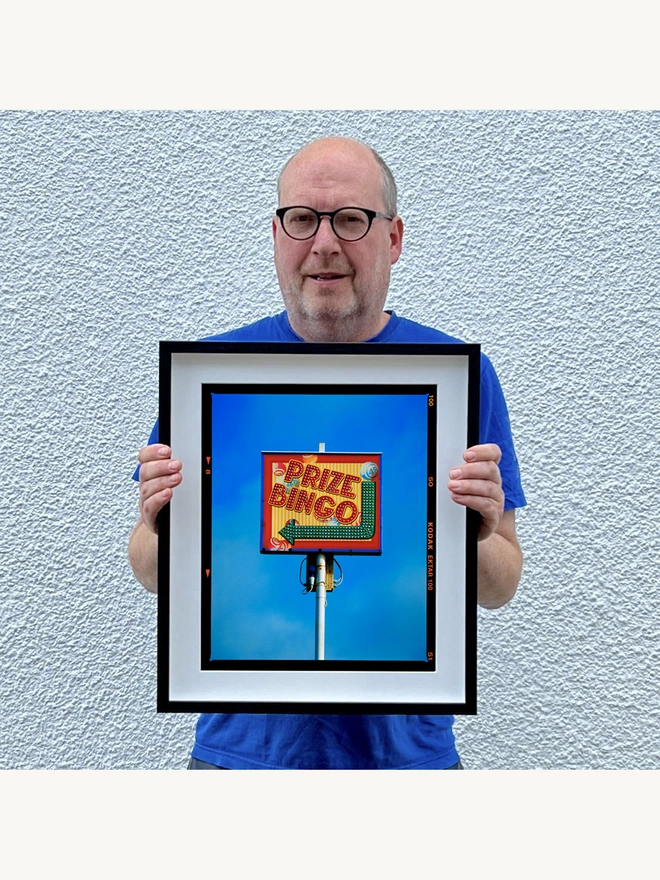 Prize Bingo Richard Heeps holding colour print framed in black