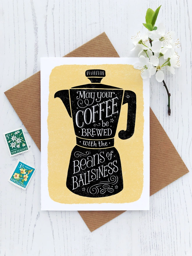 black coffee cup on yellow background with white hand lettered coffee quote inside with postage stamps and white flowers