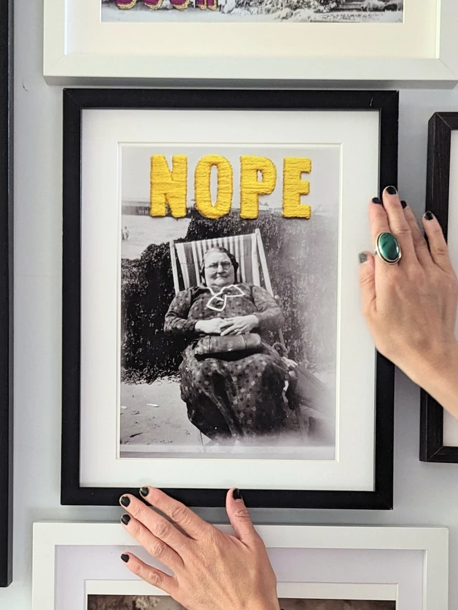  B & W photo print of woman with yellow embroidered ‘nope’ framed held on wall 