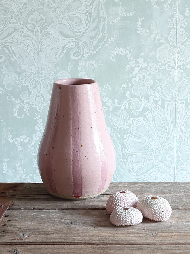 Handmade Ceramic Stoneware Large Vase in Pink Stripe sat on a wooden surface against a white and pale blue patterned wallpaper. A collection of three pink sea urchins sits next to the vase. 