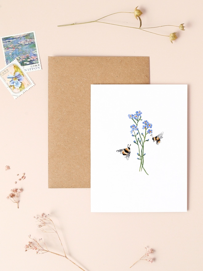 An image of a mini, rectangular white greetings card with an illustration of forget me not flowers and a couple of bumble bees. The mini greetings card is placed on top of a rectangular brown kraft envelope.