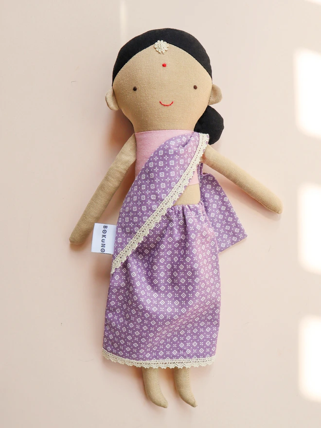 Handmade fabric doll with medium olive skin tone, wearing a lilac patterned sari with lace trim and a pink top. The doll has black hair styled in a low side bun with a small white flower accessory on her forehead, simple stitched facial features. 