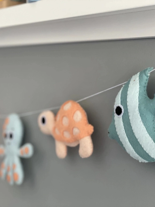 Nursery Under The Sea Garland, a sea themed garland hanging from a white shelf against a grey backdrop in a child's bedroom. 