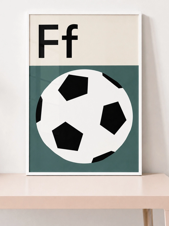 A framed print featuring the letter 'F' and a football / soccer ball on a green background, displayed on a wooden table.