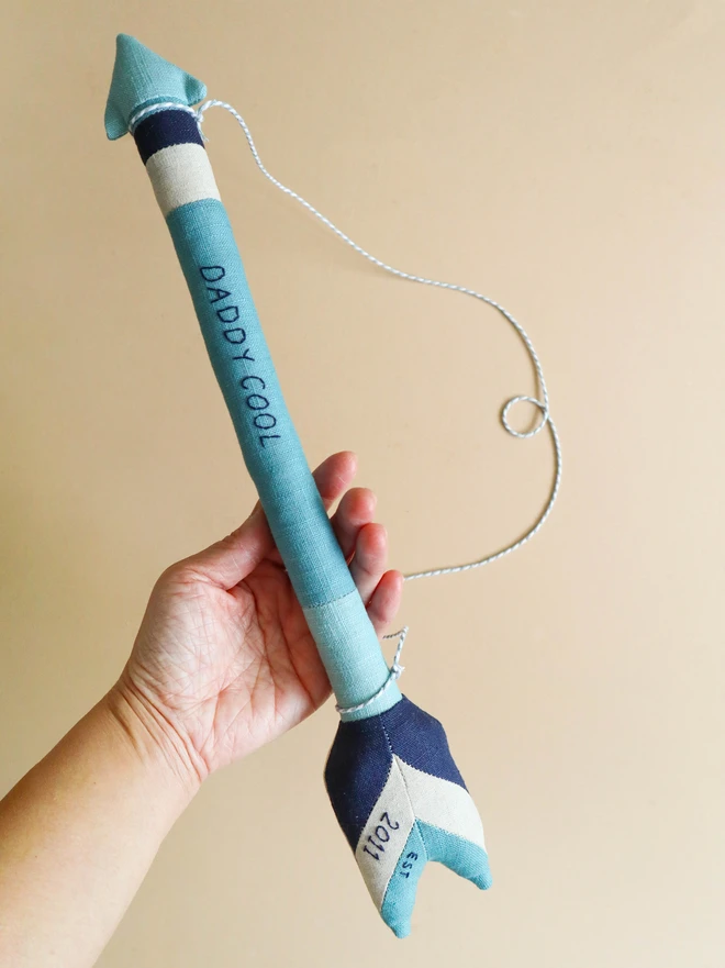 A handmade linen arrow wall hanging in shades of blue and navy, embroidered with the words “DADDY COOL” along the shaft and “2011” on the fletching. A white and grey cord is attached for hanging.
