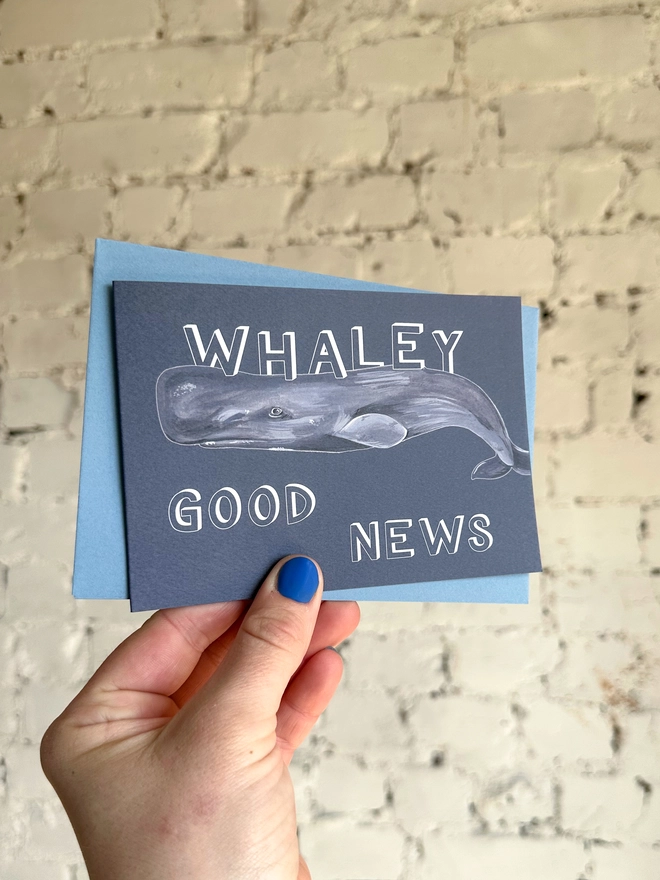 a dark grey blue greetings card featuring an illustration of a sperm whale with the text “whaley good news” with a blue envelope