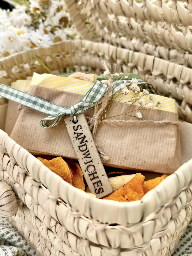 woven picnic basket holding sandwiches wrapped in brown paper that have been tied with green gingham ribbon, holding a ceramic tag embossed with 'sandwiches'.