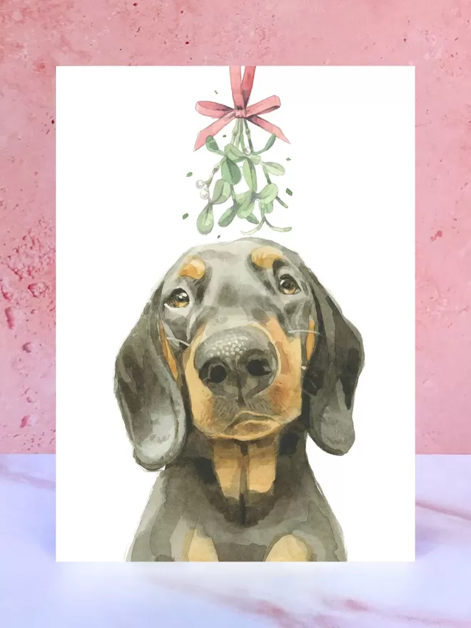 A Christmas card featuring a hand painted design of a Black And Tan Dachshund, stood upright on a marble surface.