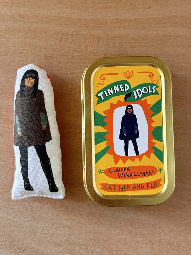 Mini fabric doll featuring an illustration of Claudia Winkleman in her Traitors cape. The doll is beside a gift tin.