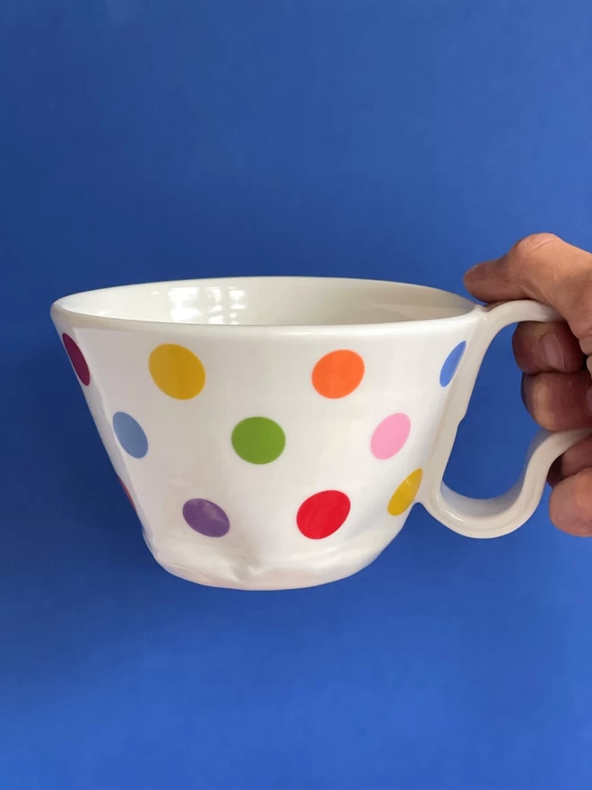 hand holding a large porcelain cup with pattern in yellow orange blue green pink purple and red polkadots on a bright blue background