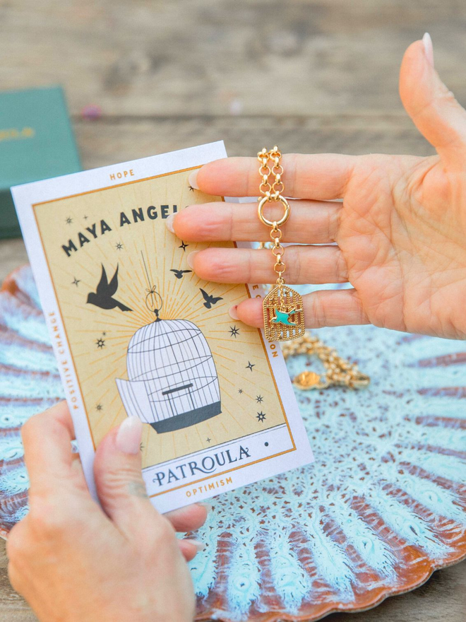 Woman's hand holding a gold belcher chain necklace with a Maya Angelou bird and gilded cage charm against a blue artisan plate and a yellow Maya Angelou card