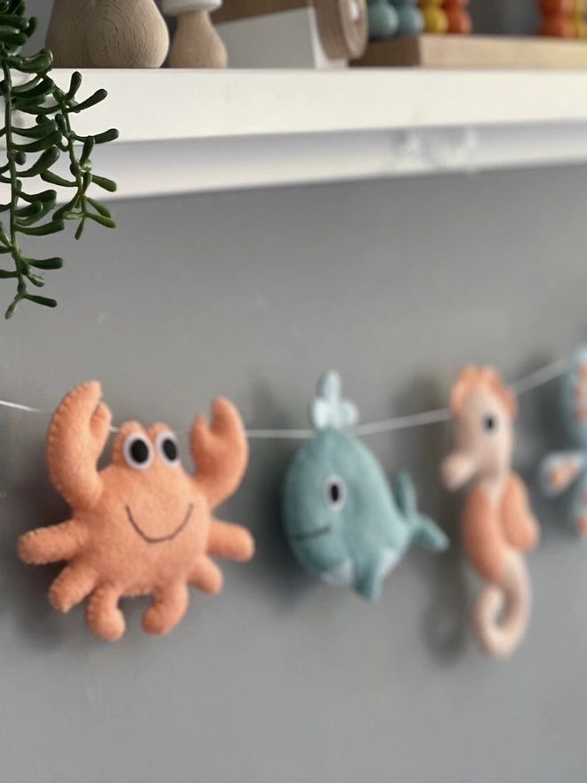 Nursery Under The Sea Garland, a sea themed garland hanging from a white shelf against a grey backdrop in a child's bedroom. 