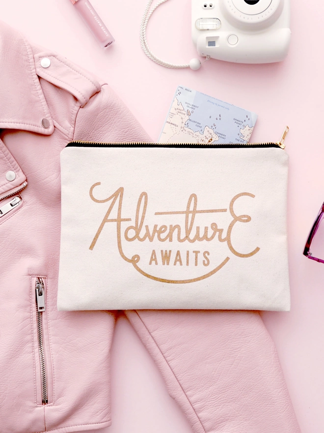 a natural canvas zip pouch with the words adventure awaits laying on a pink backdrop with a map, camera and jacket laying around