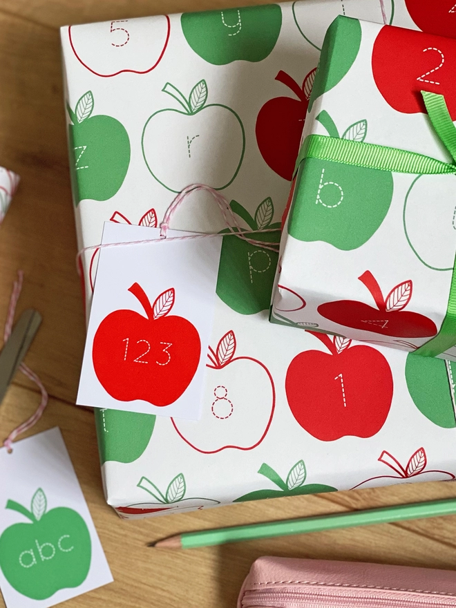 A gift wrapped in red and green apple wrapping paper, with letters and numbers inside each apple, is on a wooden surface.