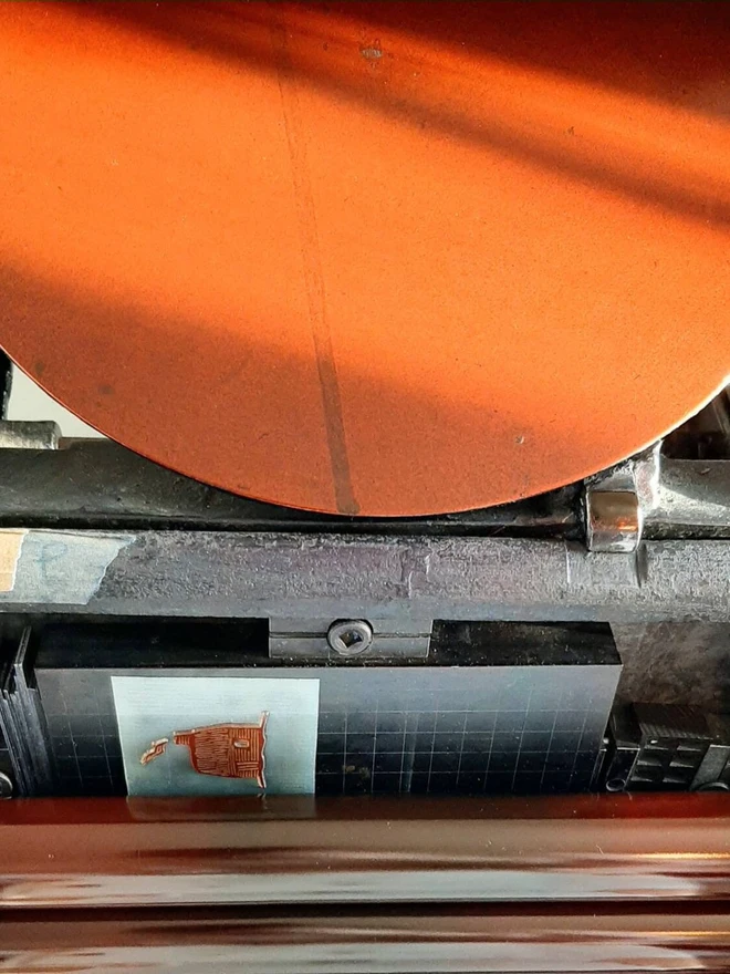 Tiny Red Cottage Letterpress Handprinted Greetings Card, a white card with a tiny red cottage design in the centre being made by a machine in a studio. 