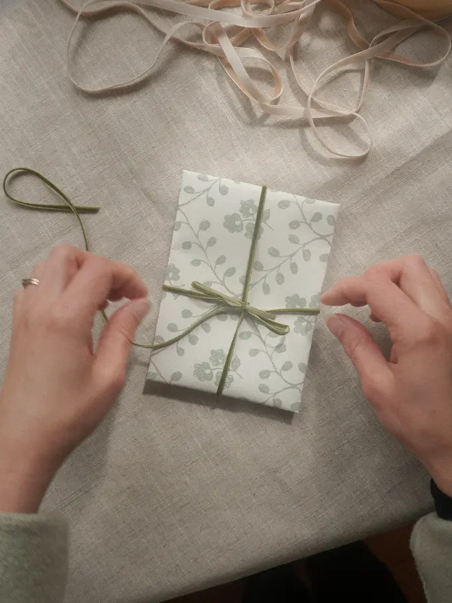 Hands wrapping a gift in botanical printed paper and olive green velvet ribbon.