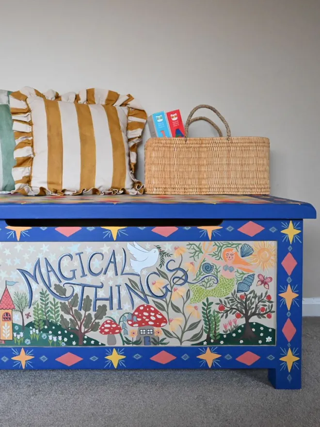 A bright blue wooden chest with a hinged lid, painted with stars and whimsical illustrations, including a castle and mushrooms, and the words "Magical Things" in the center. Two striped pillows and a woven basket sit on top.