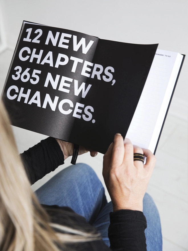 Lady holding a HELLO TIME planner open on a black quotes page reading '12 new chapters, 365 new chances.'