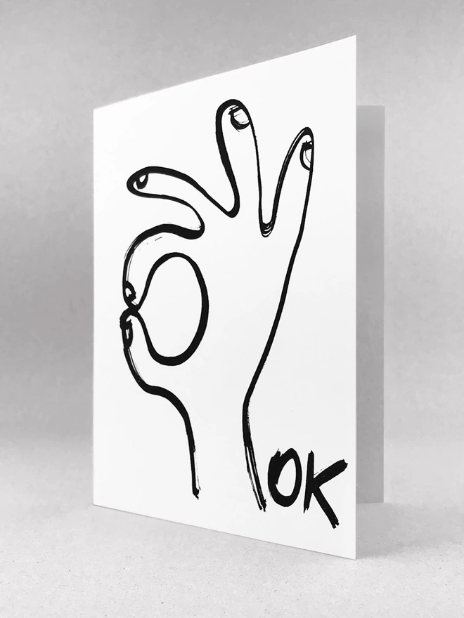 A white Ok Greetings Card with a black ink drawn hand and the word ok, slight 3/4 view made by Salty's Studio.