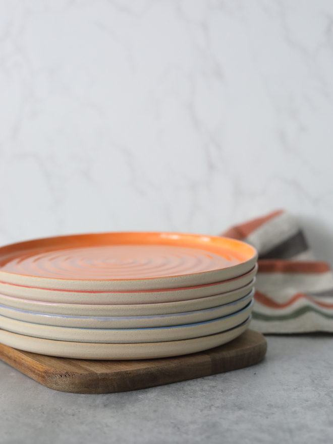 A close-up stack of handmade ceramic lunch plates on a wooden board, topped with a mandarin orange plate showing a concentric swirl texture, with coloured rim edges visible beneath — including red, lavender, and blue — styled with a striped linen cloth against a marble background