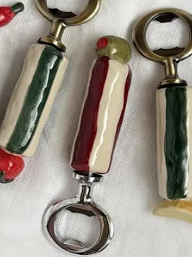 Olive, Chilli, Or Pickle Bottle Openers, a group of bottle openers on a white tablecloth. They each have a small food or fruit design. 
